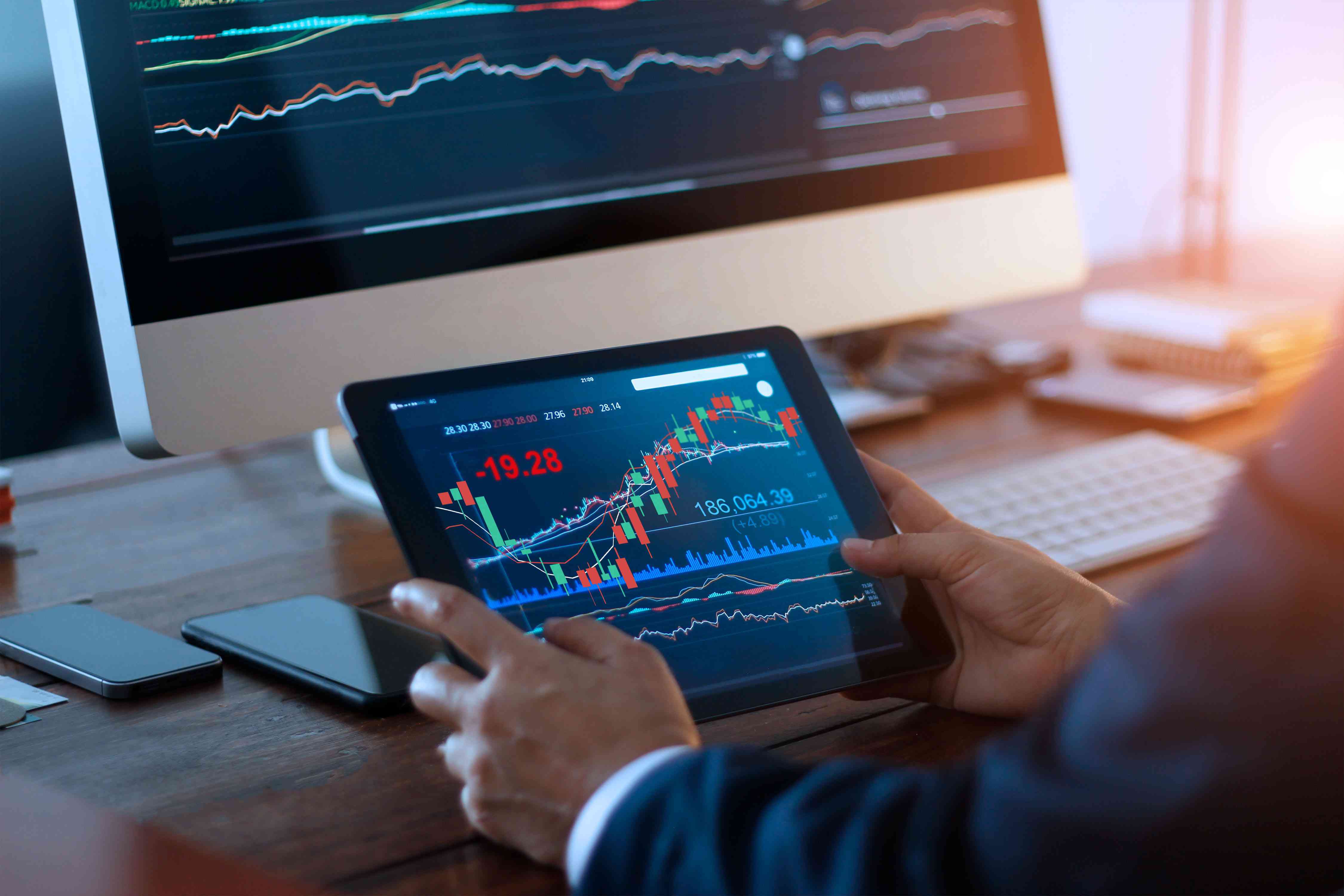 Businessman checking stock market on digital tablet and a desktop computer with stock exchange graph on screen. Financial stock market. Analyzing data in office background.