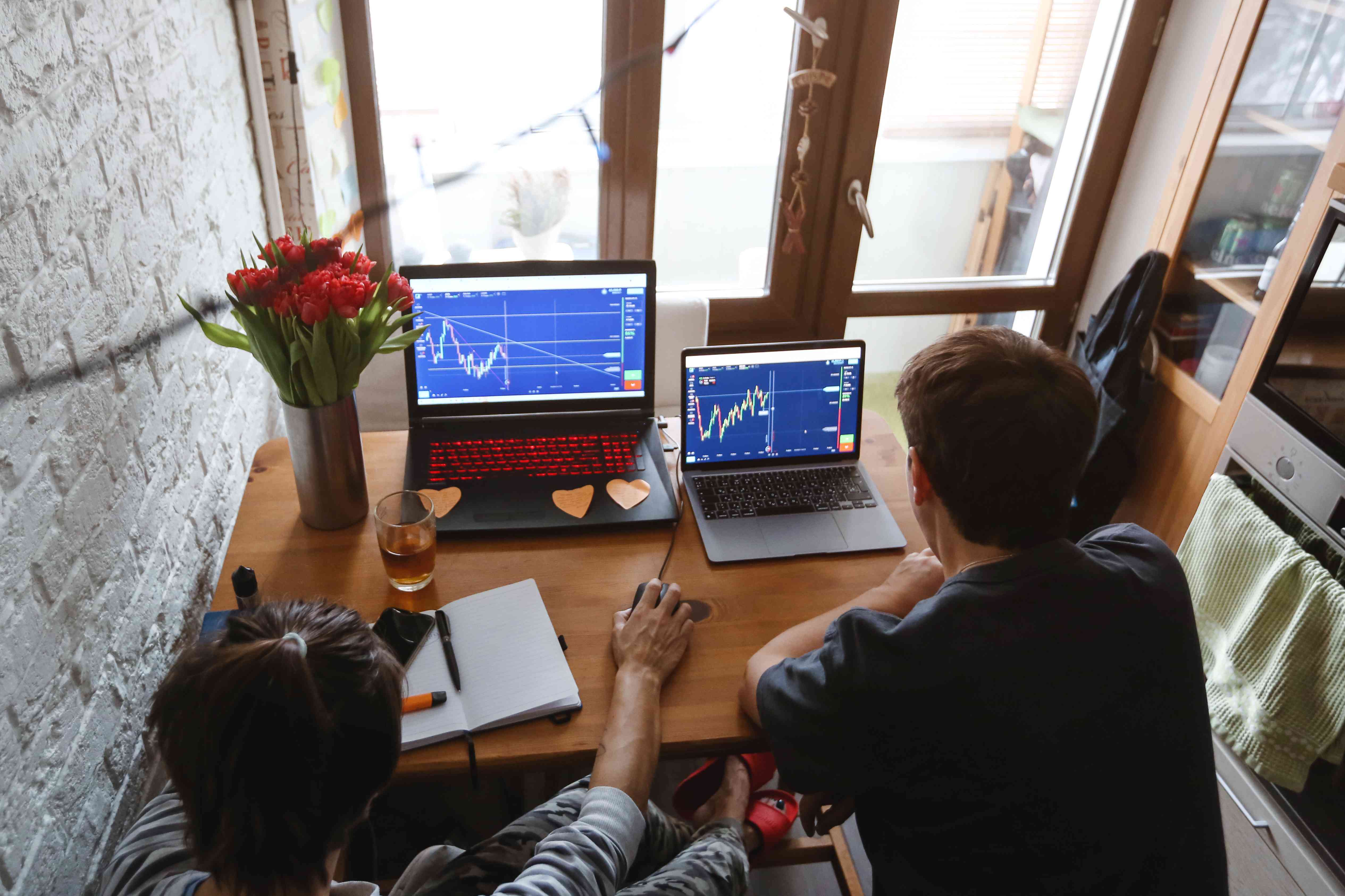 A couple of stock traders sitting at home with stock charts showing technical indicators on laptop screens, using notebook to learn about technical analysis.