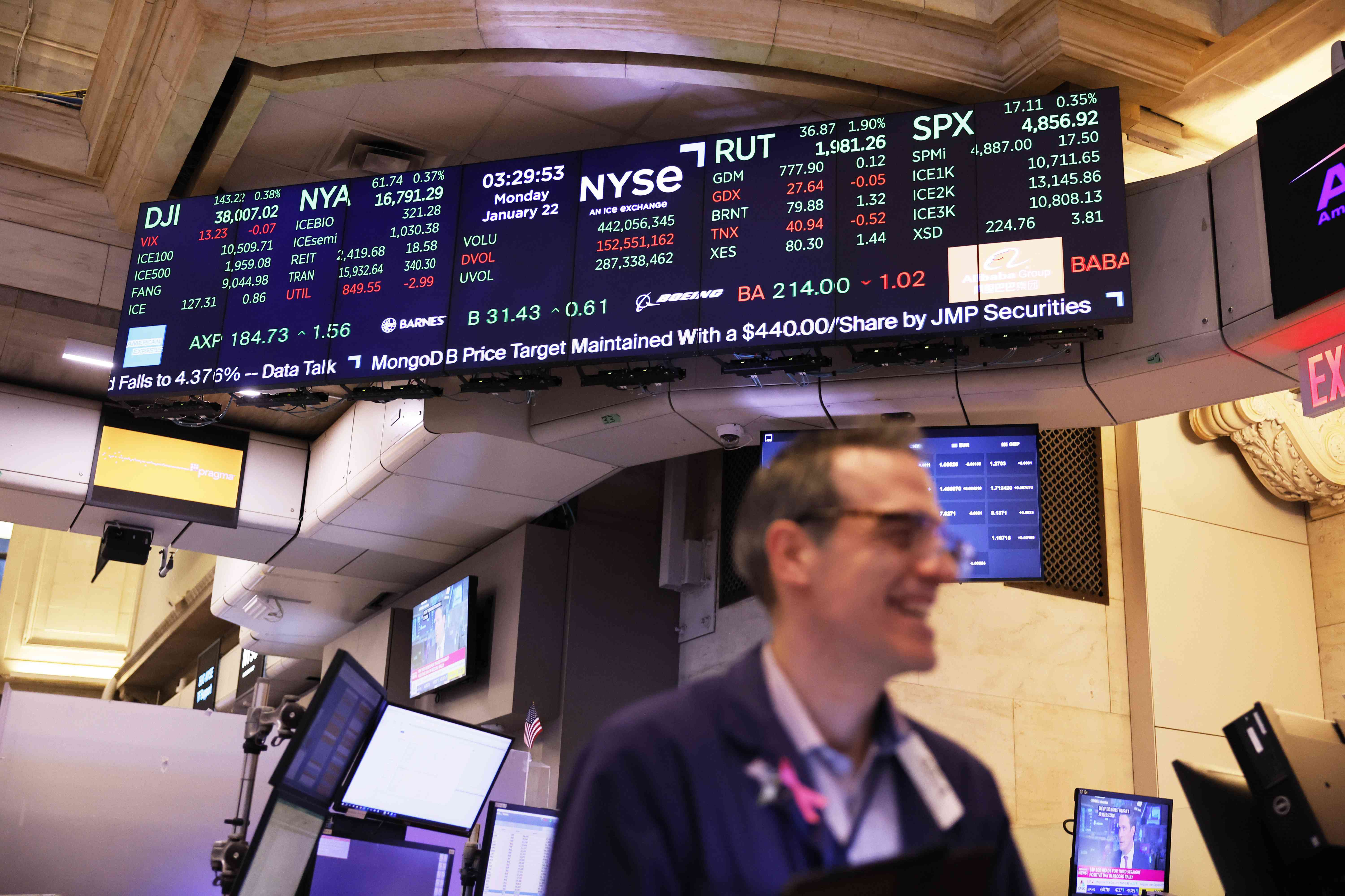 Stock market numbers are a displayed on a screen at the New York Stock Exchange, as a trader works in the foreground.