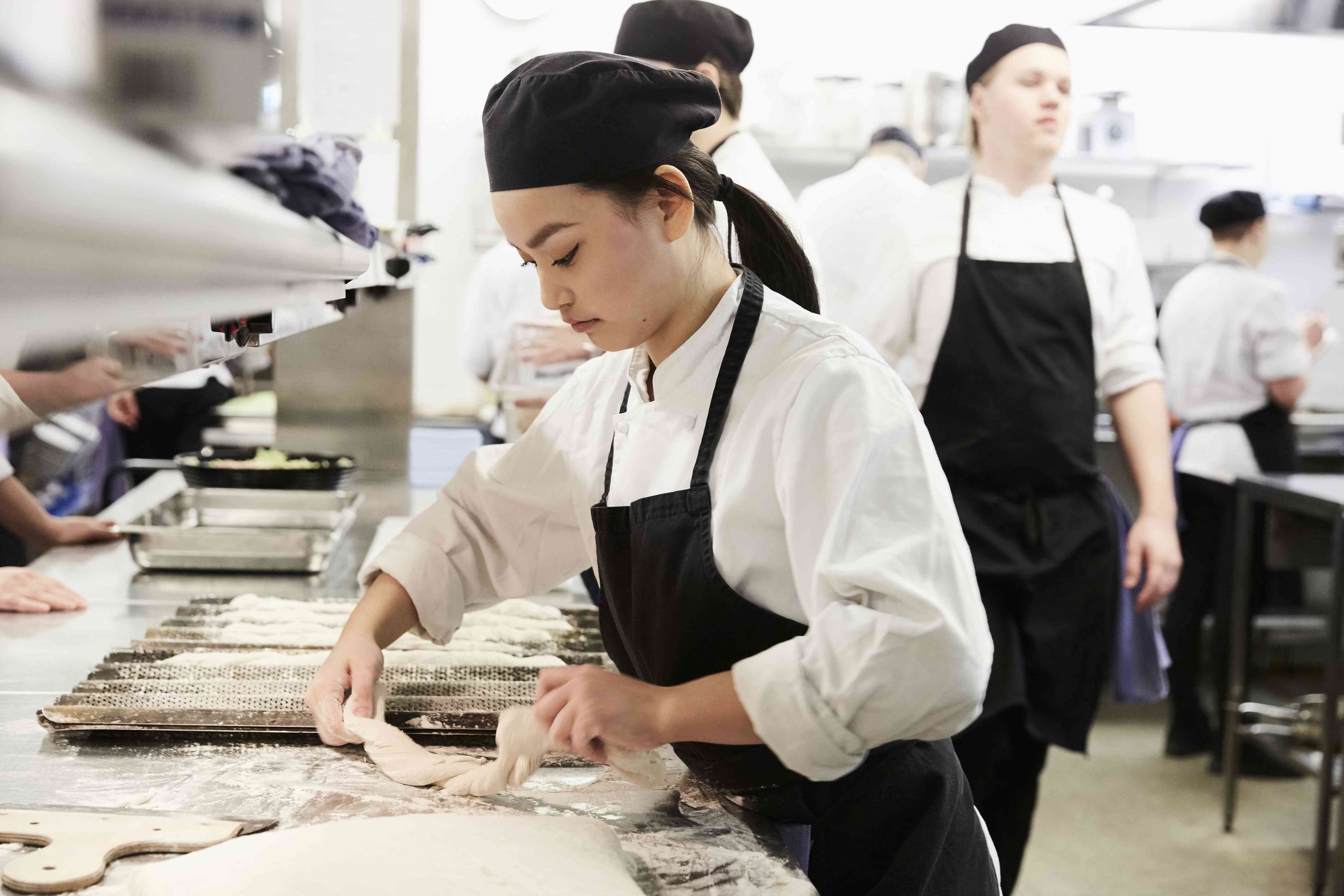 A young pastry chef works with dough on a work surface in a restaurant kitchen.