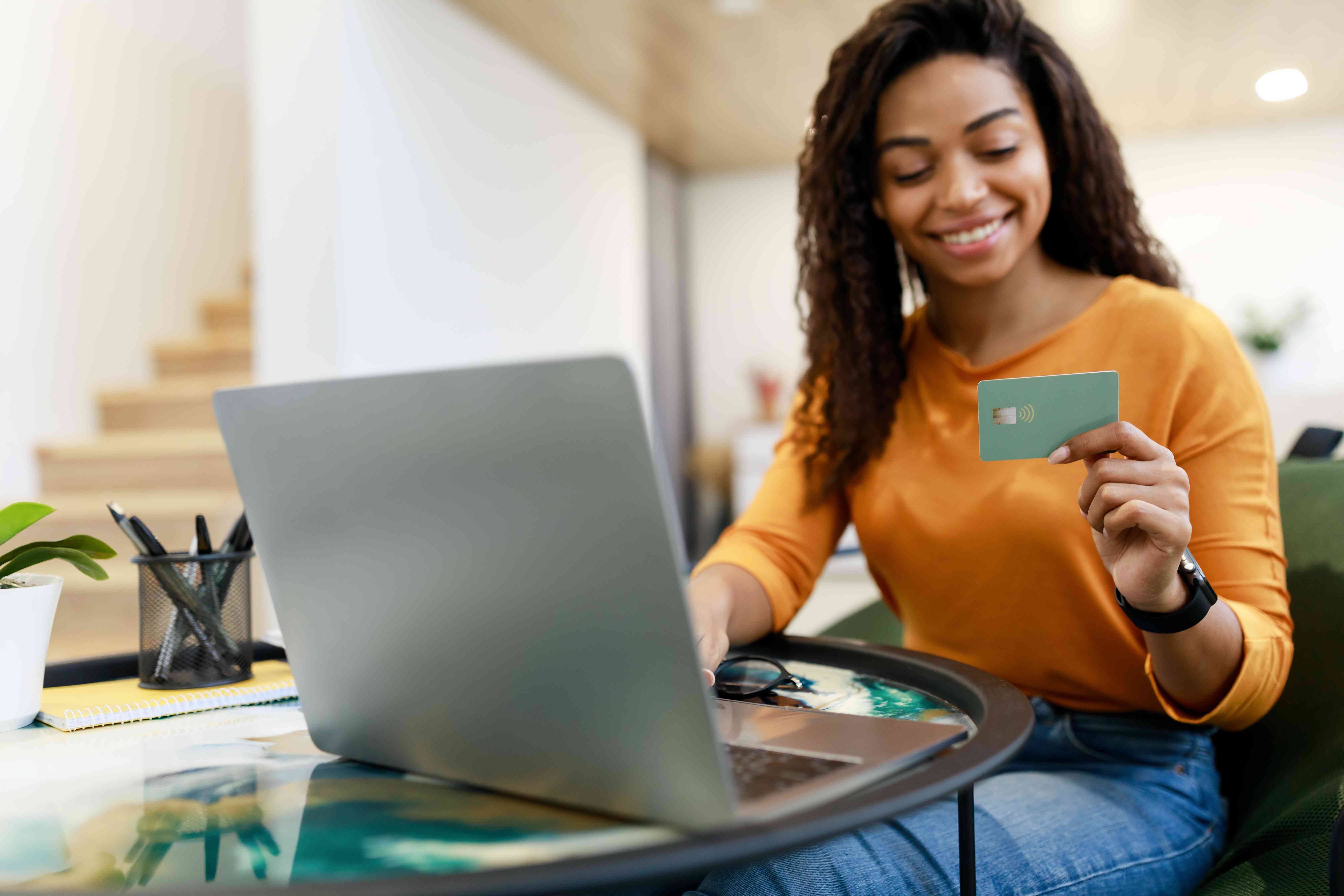 A happy woman sits a table with a laptop, smiling at a debit card in her hand.