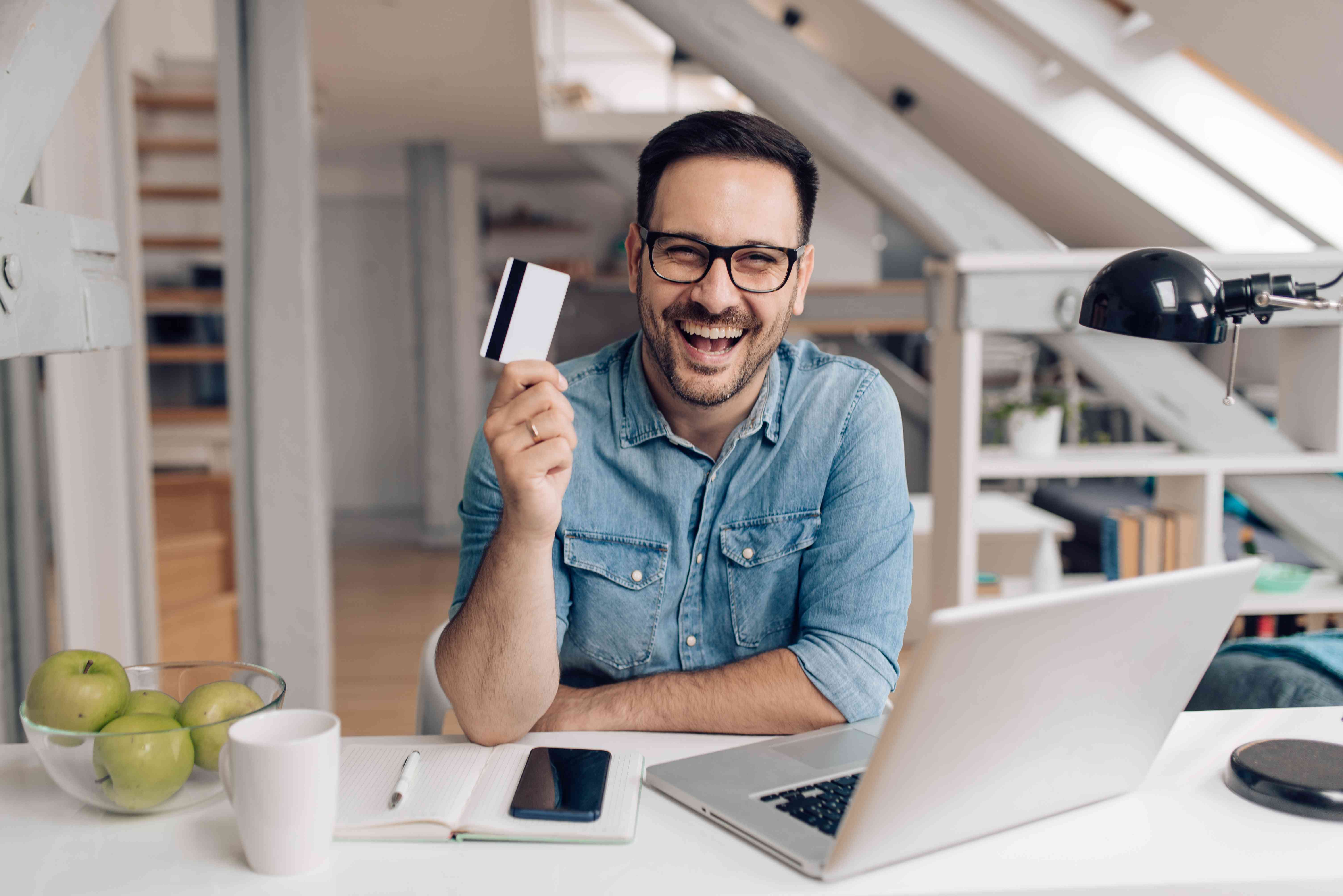 A happy young man sits at a desk with a computer, holding up a debit card.