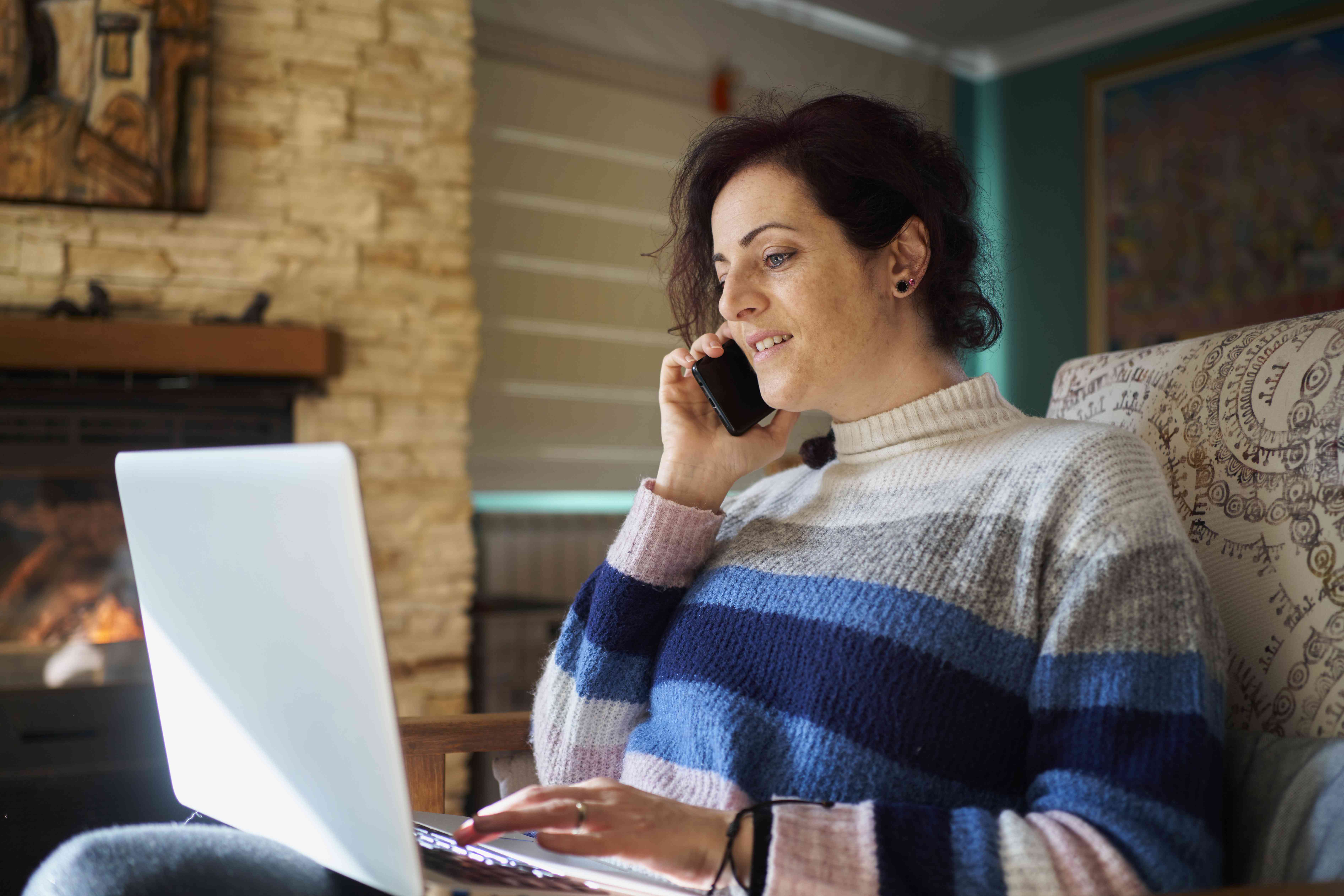 A woman sits on a chair with a laptop in her lap while talking on the phone