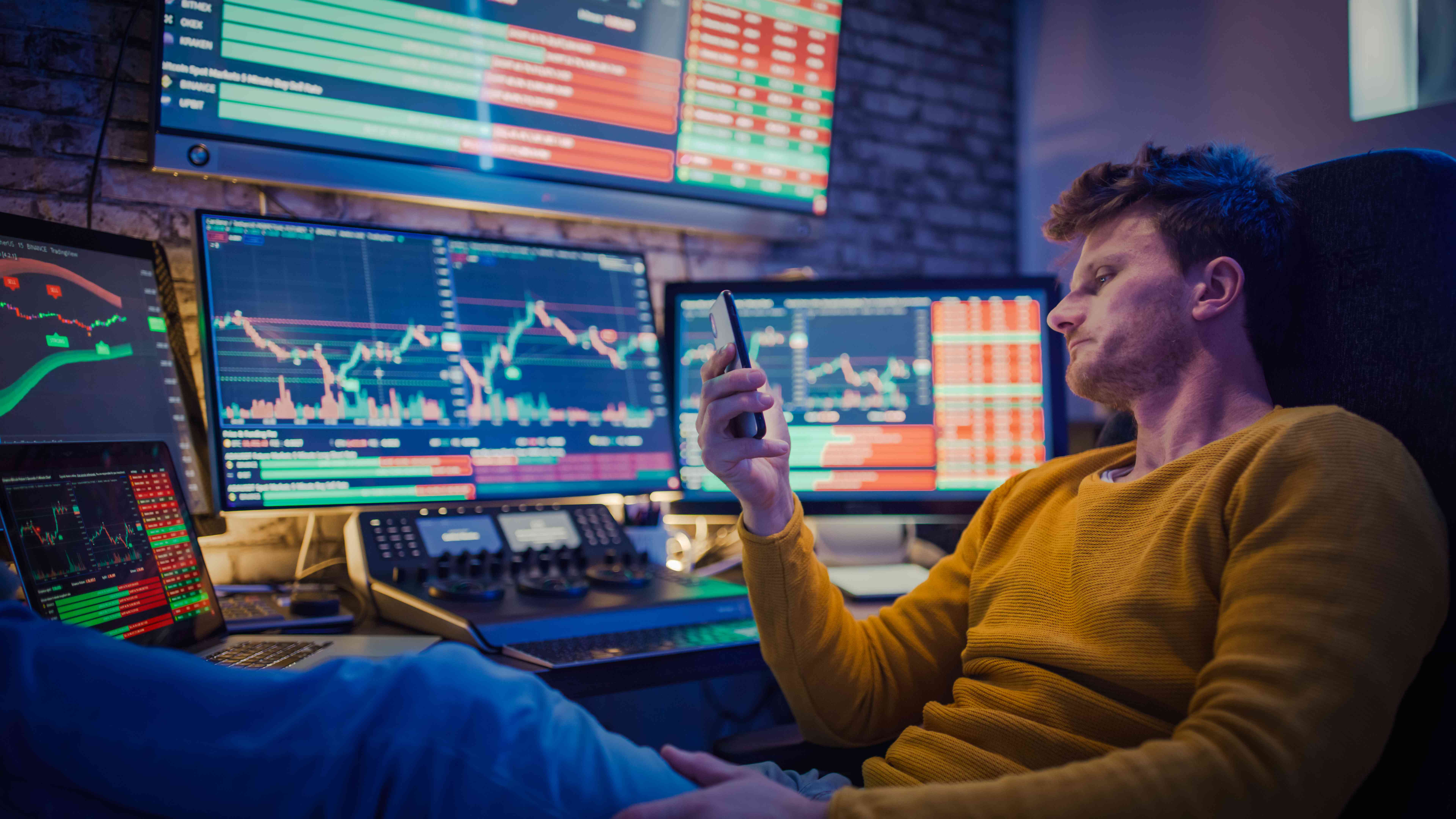 Trader looking at his phone in front of his desk with multiple monitors and programs.