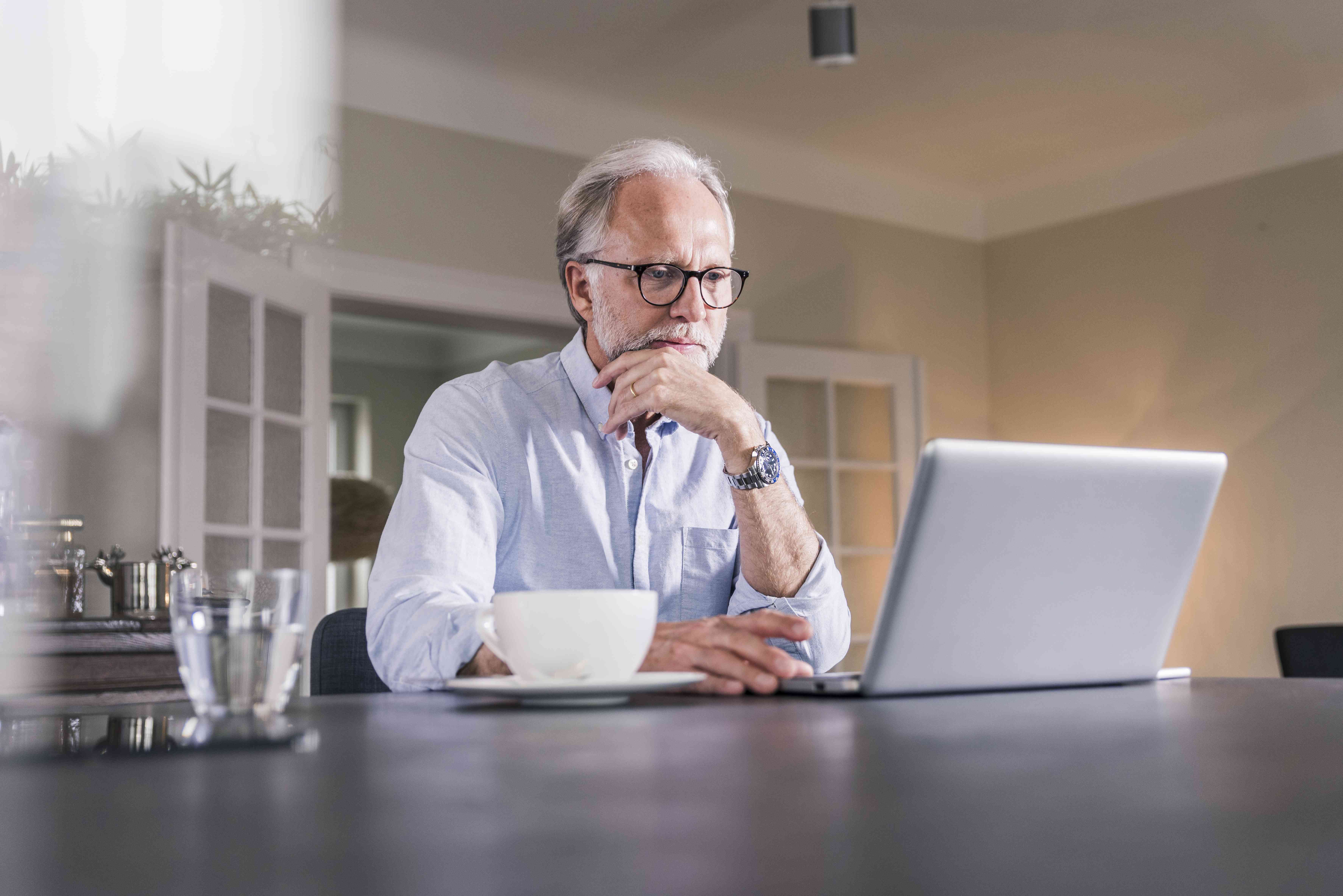 Older man sitting at his dining room table and looking intently at his laptop screen